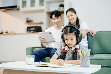 Fototapeta premium Asian child wearing headphones, studying and drawing at home, while parents enjoy coffee and tablet nearby. Family time, learning