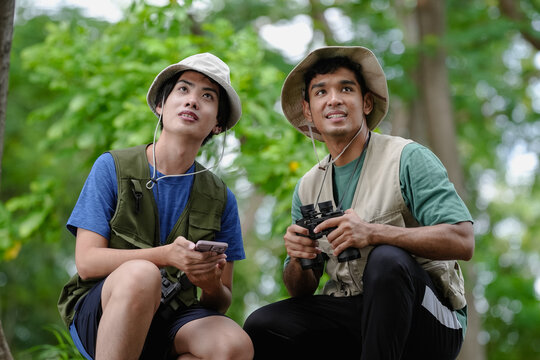 Bird watching is recreational pursuit. Young man and friend have fun with outdoors leisure activity looking up for bird in nature public park. Gay couple enjoy using binocular for bird watch adventure - Powered by Adobe