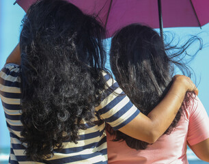 Two women with long hair are standing under a pink umbrella