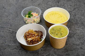 lunch soup, mashed potatoes and onion cutlet with salad in disposable dishes on a gray background