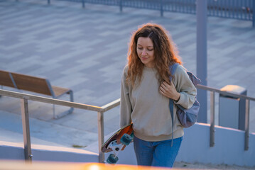 Young woman with curly hair and casual clothing walks down stairs while carrying a skateboard and...