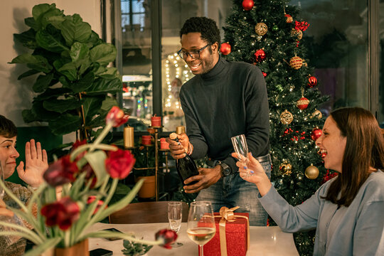 Happy multicultural couple laughing at a Christmas party celebration at home. Man opening a bottle of champagne for a festive holiday toast with friends during a New Year's Eve dinner. - Powered by Adobe