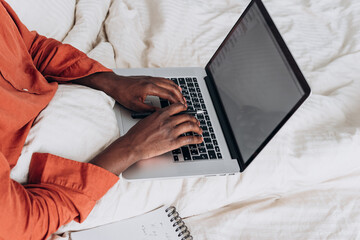 Freelancer African American is working from home, typing on a laptop while sitting in bed, with a...