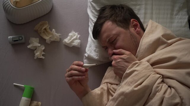 Male patient lying in bed, wrapped in blanket, experiencing severe cold symptoms, coughing while holding thermometer, surrounded by tissues and nasal spray