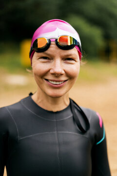 Smiling woman in a wetsuit, pink swimming cap, and goggles outdoors