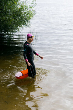 Woman in wetsuit with swimming goggles and buoy enters water on lake