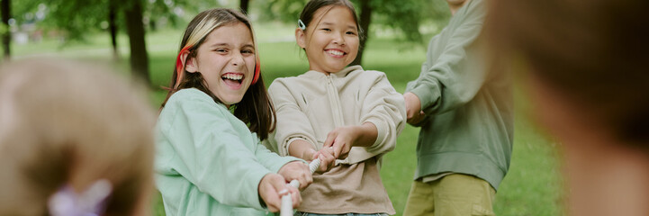 Multiethnic group of children pulling rope together outdoors in park, smiling and laughing while...