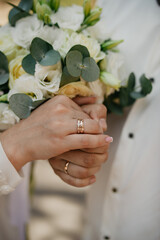 The bride in a white beaded dress next to the groom in a white shirt holding each other's hands with gold wedding rings on their fingers against the background of a bouquet of white roses