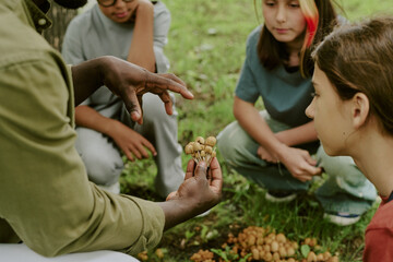 Black man teacher showing group of multiethnic children mushrooms outdoors in park, children sitting on grass attentively observing and listening during educational nature lesson