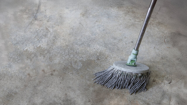 Close-up image of a round mop head cleaning an old, stained concrete floor. The image shows cleaning in progress, with visible texture and cracks on the floor surface