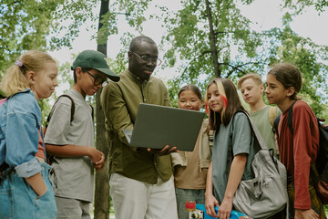 Black male teacher showing laptop to group of multiethnic children standing outdoors in park, children watching screen attentively, backpacks visible, trees in background, educational activity