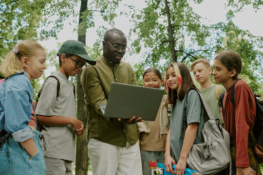Black male teacher showing laptop to group of multiethnic children standing outdoors in park, children watching screen attentively, backpacks visible, trees in background, educational activity - Powered by Adobe