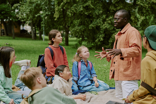 Group of diverse children sitting on grass listening to Black male teacher explaining lesson outdoors in park, children appearing engaged and attentive, backpacks visible, trees in background