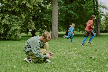 Caucasian boy tying shoelaces on grass while two children and female teacher running in background in park, group engaging in outdoor school activity surrounded by trees