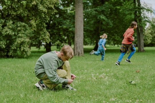 Caucasian boy tying shoelaces on grass while two children and female teacher running in background in park, group engaging in outdoor school activity surrounded by trees