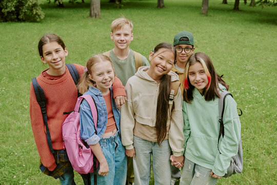 Group of diverse children standing together in park smiling at camera, multiethnic boys and girls with backpacks posing outdoors during school outing or educational activity