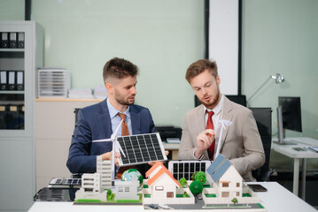 Professionals in suits discuss green energy solutions with solar panels, wind turbines, and building models in a modern office.