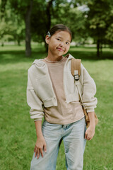 Portrait of Asian girl child standing outdoors in park wearing backpack, smiling at camera, posing...