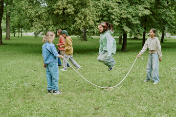 Group of multiethnic children playing jump rope while other children waiting turn in green park setting during outdoor school activity