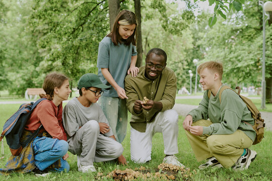 Black male young adult teacher showing mushrooms to group of diverse children and teenagers sitting and kneeling on grass in park, students observing and learning outdoors together