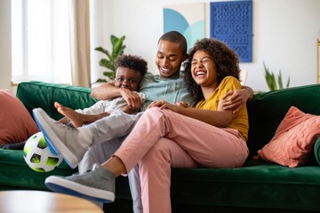 Happy african american family relaxing on couch at home with soccer ball and laughing