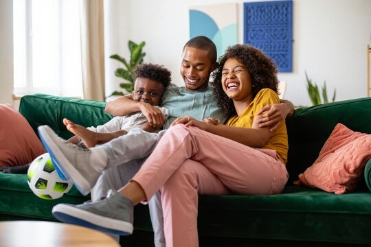 Happy african american family relaxing on couch at home with soccer ball and laughing - Powered by Adobe