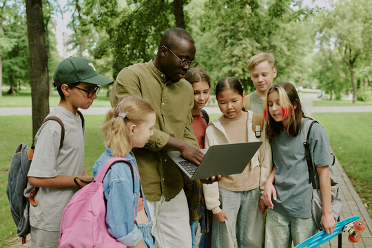 Black male teacher showing laptop to group of multiethnic children outdoors in park, children standing closely and watching screen, backpacks visible, learning together during outdoor school activity