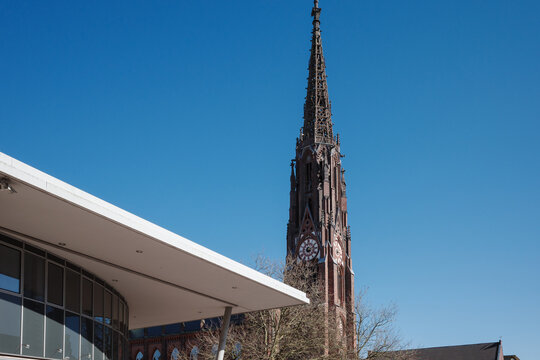 A striking architectural contrast between old and new in a German city. The tall, ornate spire of a historic Gothic Revival church stands against a clear blue sky - Powered by Adobe
