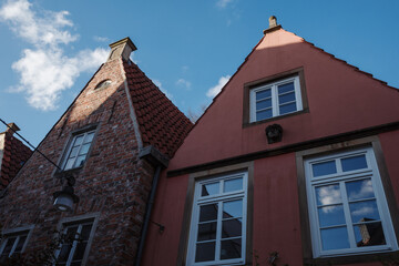 Two traditional gabled houses, one with exposed red brick and the other with a reddish-pink stucco facade, stand side by side under a bright blue sky with wispy white clouds. 