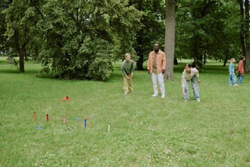 Multiethnic group of children playing outdoor ring toss game with Black male teacher supervising in park, kids focusing on activity while standing on grass surrounded by trees