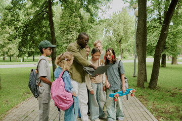 Group of diverse children and Black male teacher standing outdoors in park, teacher showing...
