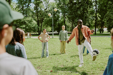Black male young adult teacher demonstrating soccer skills to group of diverse children standing in...