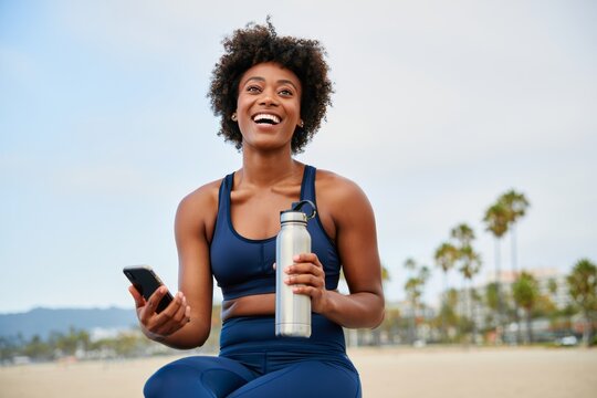 Woman athlete after workout holding phone and water bottle fitness lifestyle on beach - Powered by Adobe