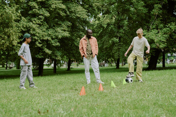 Obraz premium Diverse Boys playing soccer with cones on grass in park with young Black male teacher, engaging in outdoor activity together