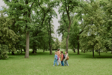 Fototapeta premium Group of multiethnic children walking with Caucasian female teacher through green park, kids carrying backpacks, teacher leading students outdoors, trees and grass surrounding group