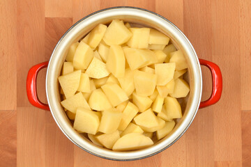 This high-angle, top-down studio shot features a pot filled with freshly peeled and roughly chopped potatoes on a wooden background, ready for cooking
