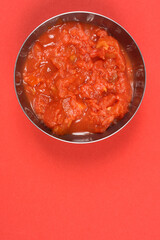 This studio shot, close-up, top-down view features a metal bowl filled with rich, chunky crushed tomatoes, prominently displayed on a vibrant red background