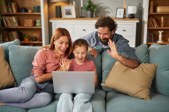 A mother, father, and daughter gather around a laptop on a couch, sharing excitement and smiles as they interact with something on the screen, enjoying quality family time.