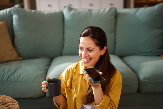 A woman with long hair wears a bright yellow shirt while sitting on a teal couch. She smiles as she holds a smartphone to her ear and enjoys a cup of coffee. - Powered by Adobe