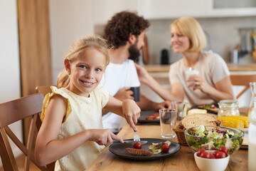 A young girl at a dining table smiles while cutting her meal. Two adults engage in conversation nearby, creating a warm family atmosphere as they share food and drinks.