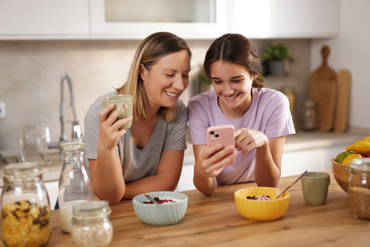 A mother holds a warm drink while her daughter shows something on her smartphone. They are sitting at a kitchen counter with bowls of cereal and fresh fruit around them.