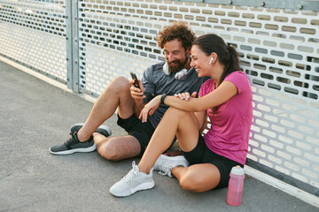 Two friends sit against a fence after working out, smiling as they review fitness stats on a...