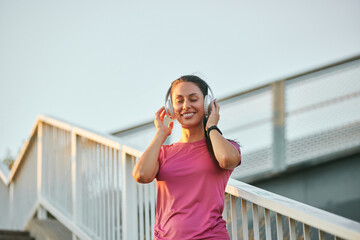 A woman in a pink athletic shirt runs up an outdoor staircase, wearing headphones and smiling. The evening sun casts a warm glow, enhancing her enjoyment of the activity.