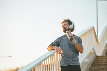 A man takes a moment to relax on a bridge after jogging, wearing headphones and holding a water bottle, surrounded by a peaceful park at sunrise, embracing a healthy lifestyle.