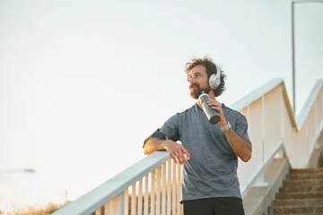 Selbstklebende Fototapeten Entspannung A man takes a moment to relax on a bridge after jogging, wearing headphones and holding a water bottle, surrounded by a peaceful park at sunrise, embracing a healthy lifestyle.  © Stockphotodirectors