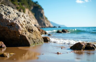 Sunlit rocky beach with gentle waves and distant cliffs under a clear blue sky