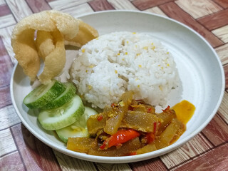 A serving of mixed rice, warm white rice accompanied by stir-fried spicy kikil with sliced red chilies and fresh cucumber on a white plate, served on a checkered wooden table.