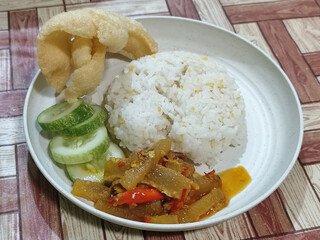 A serving of mixed rice, warm white rice accompanied by stir-fried spicy kikil with sliced red chilies and fresh cucumber on a white plate, served on a checkered wooden table.