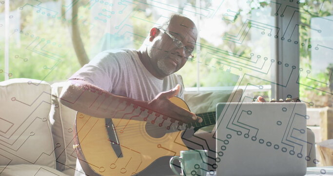 Sitting senior man wearing glasses holding guitar on sofa in studio, with laptop and coffee mug - Powered by Adobe