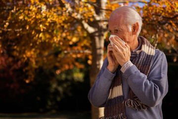 Senior man blowing nose with tissue in autumn park with yellow and orange leaves, wearing scarf